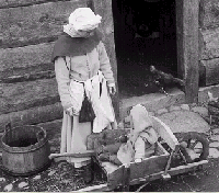 [Billede: Person iklædt gammeldags tøj. Står ved en gammeldags trillebør, hvori der sidder et barn.]