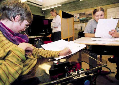 [Billede: Her ses en pige, der sidder i et klasseværelse i sin kørestol. Foran sig har hun nogle papirer liggende.]