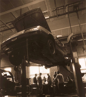[Billede: Her er vi på et autoværksted. I forgrunden ses en bil, der er hejst op mod loftet, og bagved ses en gruppe elever.]
